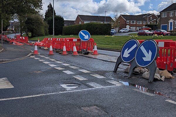 A residential street with temporary roadworks, marked by orange cones and red plastic barriers. Two blue signs with white arrows direct traffic around the obstruction. The road surface is wet with puddles, and houses, parked cars, hedges, and a partly cloudy sky are visible in the background.