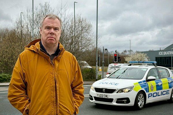A campaign-style image featuring Chris Howley standing by a roadside on a cloudy day, wearing a mustard-yellow jacket. Behind him is a marked police car stopped at traffic lights. Overlaid text reads: “Visible policing works – sign our petition to increase funding for PCSOs.